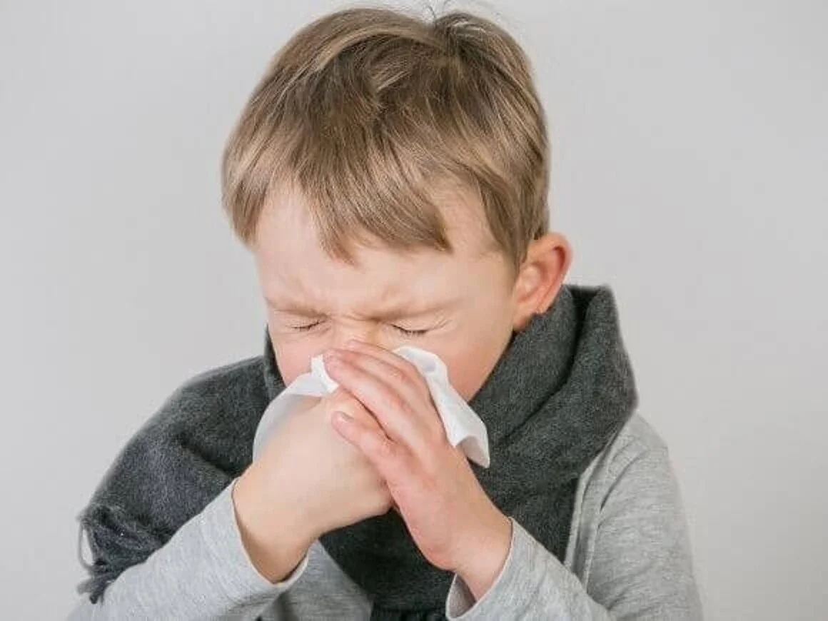 Um menino segurando um lenço de papel para assoar o nariz. Ele está com os olhos fechados e o rosto contraído, demonstrando sintomas de resfriado em crianças, como congestão nasal e coriza.